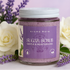 Jar of Aroma Mora sugar scrub with lavender and white flowers in the background